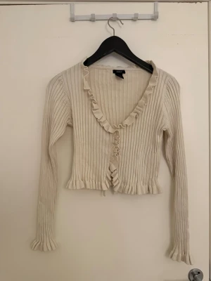 Krämvit ribbad kofta med volang - Säljer en krämvit ribbad kofta från Bik Bok med volangkant längs v-ringningen och knäppning framtill. Koftan är kortare i modellen och har långa ärmar samt fina detaljer vid ärmslut och nederkant.