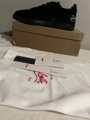 Svarta sneakers från Christian Louboutin - Snygga svarta sneakers från Christian Louboutin med diskret logga på hälen och klassisk röd detalj. Skorna har en låg siluett och är tillverkade i mocka och läder. Kommer med originalkartong, dustbag och äkthetskort.