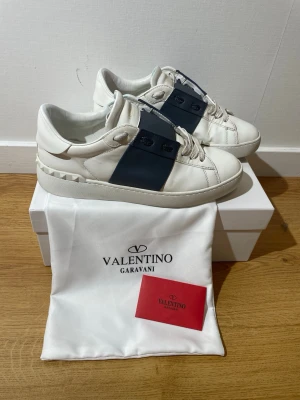 Valentino Garavani vita sneakers - Snygga vita sneakers från Valentino Garavani med bred svart rem över sidan och coola nitar på hälen. Tillverkade i slätt läder med vita snören och gummisula. Perfekta för dig som gillar exklusiv och stilren design. Den är lite smutsig med går att rengöra. hör av er för funderingar  eller bilder 😁