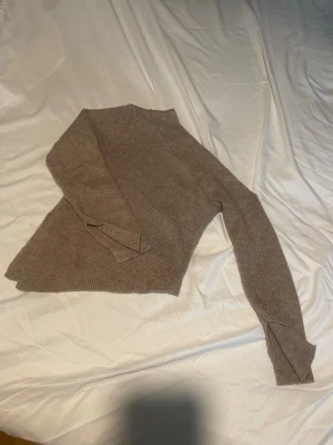 Beige ribbstickad tröja - Säljer en beige ribbstickad tröja med asymmetrisk nederkant och långa ärmar. Tröjan har en mjuk och stretchig känsla. Öppen på ena axel. 