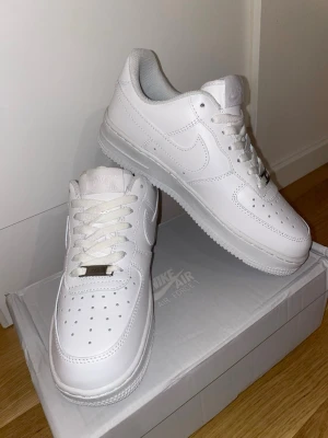 Oanvända Nike Air Force 1 vita sneakers i kartong - Helt nya och oanvända, tyvärr passade de tyvärr ej. Klassiska helvita Nike Air Force 1 sneakers i lågt utförande. Skorna har ovandel i slätt läder, perforeringar på tån för ventilation och vit sula. Swoosh-logga på sidan och metallbricka med 'AF1' på snörningen. 