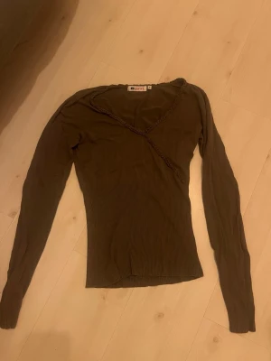 grön ribbad tröja från Gianni - Snygg grön långärmad tröja från Gianni med ribbad midja och dekorativ omlott-detalj framtill. Tröjan har smal passform och är tillverkad i ett mjukt, stretchigt material som sitter skönt på kroppen. Perfekt för dig som gillar stilrena basplagg med en twist.