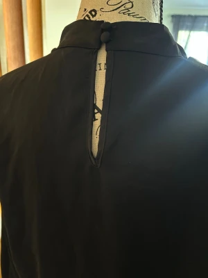 Svart oneshoulder blus med hög krage - Stilren svart oneshoulder blus med hög krage och knappstängning i nacken. Blusen har en elegant, asymmetrisk skärning och är tillverkad i ett mjukt, slätt tyg som ger ett exklusivt intryck. Perfekt för dig som vill ha en unik och trendig look.