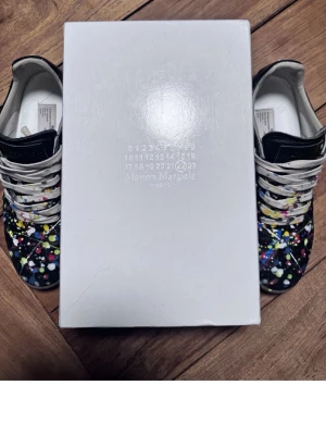 Maison Margiela sneakers med färgstänk - Maison Margiela sneakers i svart läder med unika färgstänk i gult, blått, vitt och rosa. Klassisk låg modell med vita snören och gummisula. Perfekt för dig som vill sticka ut med en kreativ och trendig look.