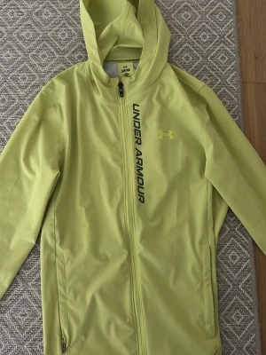 Gul vindjacka från Under Armour - Sportig gul vindjacka från Under Armour med huva och hel dragkedja framtill. Jackan har svart logga och text längs bröstet samt två sidofickor. Perfekt för dig som gillar en clean och funktionell stil.