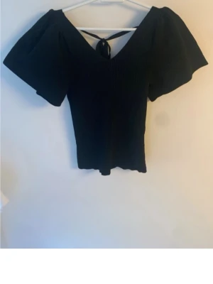 Svart ribbad topp med puffärm - Snygg svart ribbad topp med v-ringning och puffiga korta ärmar. Toppen har en knytning i ryggen och sitter figurnära över kroppen. Perfekt för dig som gillar en stilren men ändå trendig look.
