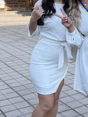 Vit omlottstudentklänning med knytband - Snygg vit kortklänning med omlottdesign och knytband i midjan. Klänningen har trekvartsärm med lätt puff och slits på sidan för en trendig look. Perfekt för dig som vill ha en stilren och modern outfit.