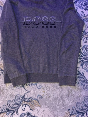 Grå sweatshirt från Hugo Boss - Snygg grå sweatshirt från Hugo Boss med stort BOSS-tryck på bröstet. Tröjan har rund halsringning, långa ärmar och ribbade muddar vid ärmslut och nederkant. Perfekt för en avslappnad och stilren look.