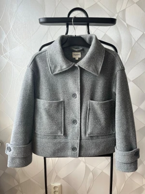 Grå ullkappa från Kappahl - Stilren grå kappa från KappAhl i ullmix med rak passform. Kappan har stora utanpåfickor, bred krage och knäppning med fyra knappar framtill. Ärmarna har dekorativa slejfar vid mudden för en extra detalj. Diskuterar gärna pris!✨