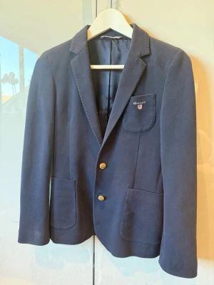 Mörkblå kavaj från Gant - Stilren mörkblå kavaj från Gant med två guldknappar framtill, klassisk krage och tre fickor. Kavajen har en broderad logotyp på bröstfickan och är tillverkad i ett mjukt tyg som ger en dressad men avslappnad look.