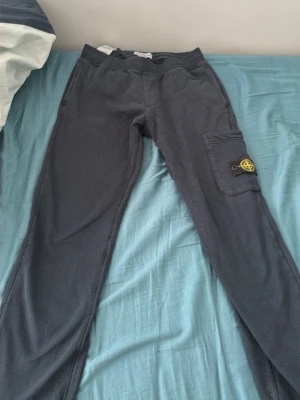Mörkgrå mjukisbyxor från Stone Island - Säljer ett par mörkgrå mjukisbyxor från Stone Island med klassisk patch på benfickan. Byxorna har resår i midjan och en praktisk sidoficka med lock. Perfekta för chill dagar och streetwear-stil.