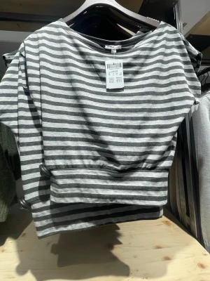 Randig grå och vit topp - Säljer en randig topp i grått och vitt med bred halsringning och avslappnad passform. Toppen har korta ärmar och är tillverkad i mjuk bomull, perfekt för en chill och enkel stil.