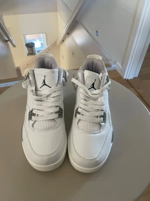 Vita Air Jordan 4 Retro sneakers - Snygga vita Air Jordan 4 Retro sneakers med grå detaljer och svart Jumpman-logga på plösen. Skorna har meshpaneler, plastdetaljer och en synlig Air-enhet i sulan. Perfekta för dig som gillar klassisk streetwear och vill ha en ikonisk look.