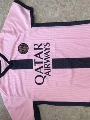 Rosa PSG matchtröja med svarta detaljer - Säljer en rosa PSG matchtröja med svarta detaljer och Qatar Airways-tryck på bröstet. Tröjan har klubbmärke på vänster sida, svarta ränder längs sidorna och runt halsen. Tillverkad i lätt och ventilerande material, perfekt för fotbollsträning eller match.