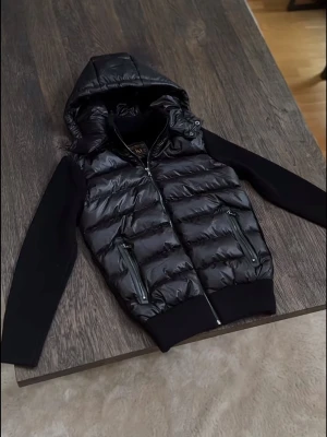 Marcél Classic 02 Hybridjacka – Svart | Avtagbar Luva | Ny Med Prislapp - Ny med prislapp — Marcél Classic 02 i helsvart med quiltad pufferkropp och stickade ärmar. Avtagbar luva, lyxiga microfleecefickor och äkta läderfickor — premium detaljer som verkligen syns och känns. Tidlös design som passar till allt. Perfekt för höst/vinter och kyliga vårdagar. Säljer direkt för ett bättre pris än butik. 📦 Spårad frakt, väl förpackad.