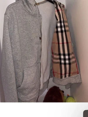 Grå hoodie med Burberry-inspirerat - Säljer en grå hoodie med dragkedja och fickor framtill. Insidan har ett klassiskt Burberry-inspirerat rutmönstrat foder i beige, svart, vitt och rött. Hoodien har huva och är tillverkad i mjukt sweatshirtmaterial. Perfekt för en chill och stilren look.