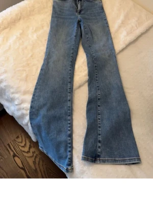 Blå bootcut jeans med hög midja - Säljer ett par klassiska blå bootcut jeans med hög midja. Jeansen har fem fickor, bälteshällor och är tillverkade i ett mjukt denimtyg. Perfekta för dig som gillar en retroinspirerad look och vill ha ett par jeans som sitter snyggt över höfterna och släpper vid benen.