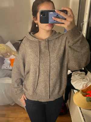 Beige stickad hoodie med huva - Mysig beige stickad hoodie med huva och dragsko. Tröjan har avslappnad passform, ribbade muddar vid ärmslut och nederkant samt långa ärmar. Perfekt för chill dagar och enkel att matcha med jeans eller leggings.