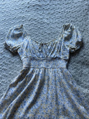 Blommig kort klänning - En mycket söt kortklänning med ljusblått och gult blommönster, puffärmar och resår i midjan. Klänningen är från Hollister och har en urringning med knyt framtill. Den är tillverkad i ett mjukt bomullstyg och har en fin liten rosett framtill. Den är i stolek S men passar även XS och M på grund av att den har resår i midjan.