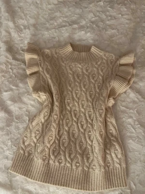 Beige stickad ärmlös tröja med volang - Supersöt beige stickad tröja med volangdetaljer vid ärmhålen och grovt kabelstickat mönster. Tröjan har ribbad hals, nederkant och ärmslut. Perfekt att styla över en skjorta eller långärmad topp för en trendig lager-på-lager-look.