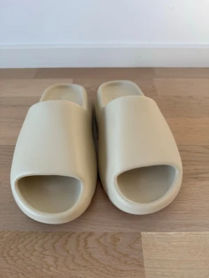 Beige Adidas tofflor - Säljer ett par stilrena beige tofflor från Adidas. De har en chunky design med öppen tå och är gjorda i ett mjukt, skummaterial som ger skön komfort. Perfekta för att glida runt hemma eller på stranden. Adidas-logga syns diskret på innersulan.