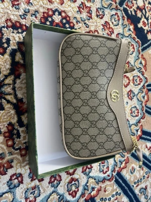 Beige axelremsväska från Gucci - Snygg beige handväska från Gucci med klassiskt GG-mönster över hela väskan. Väskan har guldfärgade detaljer, kedjerem och en elegant form. Tillverkad i canvas med läderdetaljer och stängs med dragkedja. Perfekt för dig som gillar lyxig och tidlös design.