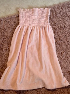 Ljusrosa morgonrock Juicy Couture - Supersöt ljusrosa morgonrock från Juicy Couture i mjuk frotté. Axelbandslös modell med smock upptill som ger en skön och avslappnad passform. Perfekt för att chilla hemma eller efter duschen. Klassisk Juicy-stil med lyxig känsla.