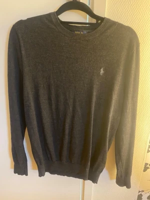 Mörkgrå merinoullströja Polo Ralph Lauren - Mörkgrå stickad tröja från Polo Ralph Lauren i slim fit-modell. Tillverkad i tvättbar merinoull med rund halsringning och diskret broderad logga på bröstet. Perfekt för en stilren och klassisk look. Storlek M passar S. Mycket bra skick förutom en lagning vid sidan av magen, syns ej vid användning, syns endast om man kollar noga 