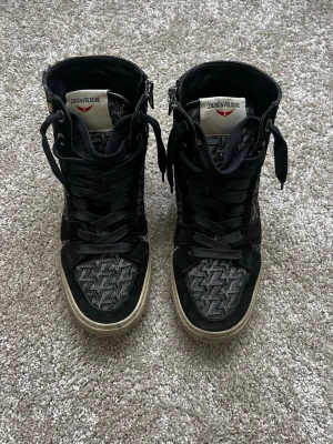 Zadig & Voltaire svarta high top sneakers - Snygga high top sneakers från Zadig & Voltaire i svart med mönstrad textil och svarta mockadetaljer. Skorna har en guldfärgad blixtdetalj på sidan och svart snörning. Perfekt för dig som vill sticka ut med en cool och unik stil. De är ganska slitna på insidan av hälen men funkar fortfarande som vanligt!