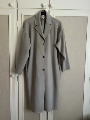 Beige kappa - Minimalistisk ljusgrå kappa med rak passform och stora slag. Kappan har tre knappar framtill, dolda sidofickor och är tillverkad i ett mjukt tyg som ger en clean look. Perfekt för dig som gillar stilrena och tidlösa plagg.