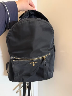 Svart ryggsäck från Michael Kors - Stilren svart ryggsäck från Michael Kors med guldfärgade detaljer och dragkedja framtill. Tillverkad i slitstark polyester med justerbara axelband och praktiskt ytterfack. Perfekt för dig som vill ha en snygg och enkel väska till vardagen. Den är i jättebra skicka men det enda med den är att den ena dragfliken inte är original.