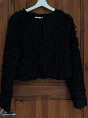 Svart textured cardiganjacka - Säljer en svart cardiganjacka med unik 3D-textur och blommiga detaljer. Jackan har rund halsringning, långa ärmar och är tillverkad i ett mjukt tyg som ger en cool och trendig look. Perfekt för dig som vill sticka ut med din outfit. Använd en gång 💞