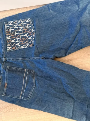 Blå jeans från NASA HSLL med tryck - Säljer ett par blå jeans från NASA HSLL med unikt tryck på bakfickan där det står 'NASA HSLL' i olika färger. Klassisk femficksmodell i denim med kontrastsömmar och patch med logga bak i midjan. Streetstyle