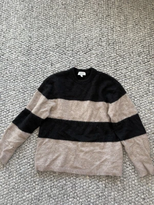 Randig stickad tröja & Other Stories - Stickad tröja från & Other Stories med breda svarta och beige ränder. Tröjan har rund halsringning och långa ärmar. Mjuk och skön kvalitet, perfekt för kyligare dagar. Enkel men snygg design som passar till det mesta.