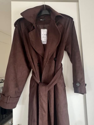Mörkbrun trenchcoat från Gina Tricot - Stilren mörkbrun trenchcoat från Gina Tricot med knytskärp i midjan. Tillverkad i ett mjukt, mockaliknande material som ger en lyxig känsla. Nypris 1099kr, säljer den då den aldrig kom till användning och inte riktigt passar min stil. Prislapp finns kvar! Helt ny som sagt☺️Även slutsåld på Gina tricot.  Kan gå ner i pris vid snabb affär!💕