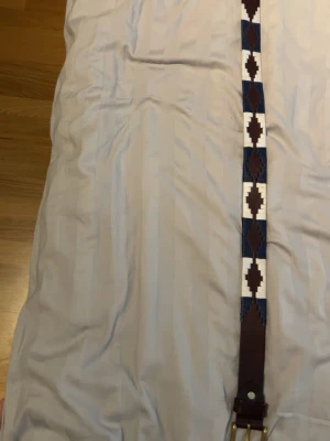 Brunt och blått mönstrat läderbälte - Snyggt bälte i brunt läder med geometriskt mönster i blått och vitt. Bältet har en klassisk metallspänne och detaljer som ger en unik look. Perfekt för att lyfta vilken outfit som helst.