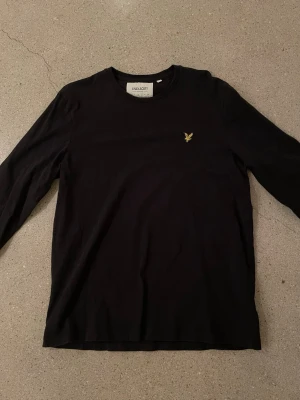 Svart långärmad topp från Lyle & Scott - Klassisk svart långärmad topp från Lyle & Scott med rund halsringning och den ikoniska gula logon broderad på bröstet. Tillverkad i mjuk bomull för en skön känsla. Perfekt till jeans för en clean look.