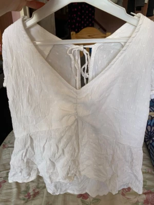 Vit blus med knyt där bak och volang - Söt vit blus med v-ringning, knyt i ryggen och volang nedtill. Blusen har korta puffärmar och är tillverkad i ett lätt, strukturerat bomullstyg med diskreta prickar. Perfekt för en somrig look och enkel att matcha med jeans eller kjol.