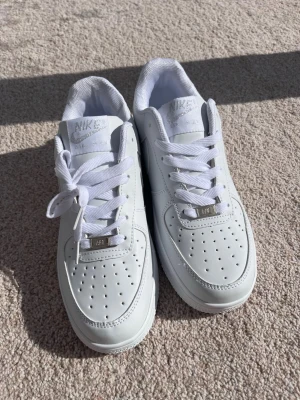Nike Air Force 1 vita sneakers - Klassiska vita Nike Air Force 1 sneakers i lågt utförande. Skorna har ovandel i slätt läder, perforerade detaljer på tån och vita skosnören. Nike-logga syns både på plösen och hälen. Perfekta för en clean och stilren look.