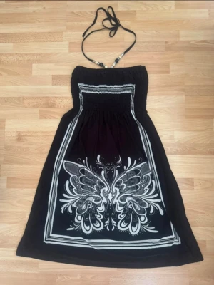 Svart långklänning med fjärilsmönster - Cool svart långklänning med vitt fjärilsmönster och smock upptill. Klänningen har halterneck med pärldetaljer och ett flowigt fall. Perfekt för dig som vill sticka ut med en unik design och snygga detaljer. Använd en gång.