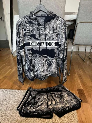 Fin dress dam  - Snyggt svartvitt pyjamas-set från Dior med hoodie och shorts. Setet har ett detaljerat mönster med djur och naturmotiv samt tydlig logga över bröstet. Hoodien har dragsko och shorts med resår i midjan. Perfekt för chill dagar skriv för pris osv 