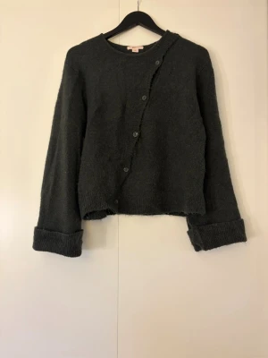 Mörkgrå asymmetrisk kofta - Mysig mörkgrön tröja med knappar och stora uppvikta ärmslut. Koftan är stickad och har en avslappnad passform.