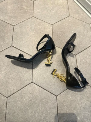 Svarta YSL pumps med gulddetalj - Säljer ett par svarta pumps från Saint Laurent med öppen tå och justerbar ankelrem. Klackarna är i guld med ikoniska YSL-loggan som design. Skorna är i lackat läder och har en elegant, modern siluett som verkligen sticker ut.
