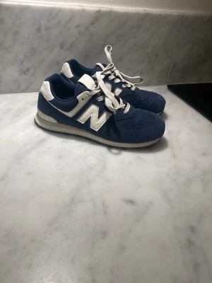 Blå New Balance sneakers - Snygga blå och vita sneakers från New Balance med klassisk N-logga på sidan. Skorna har vita snören, vit sula och detaljer i mocka och textil. Perfekta för dig som gillar sportig och stilren look.