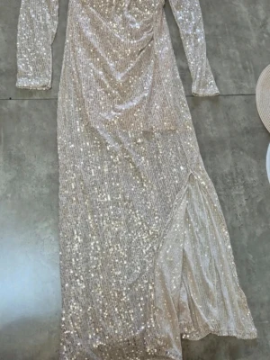 Glittrig långklänning med slits - Säljer en glittrig långklänning i ljus ton med långa ärmar och hög slits. Klänningen har ett omlottliknande fall framtill och är täckt av paljetter som ger ett riktigt festligt intryck. Perfekt för dig som vill sticka ut på nästa event!