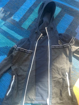 Marinblå hoodie från Maya Delorez - Snygg marinblå hoodie från Maya Delorez med dragkedja och huva. Tröjan har vita detaljer, två fickor med dragkedja och logga på bröstet. Tillverkad i mjukt funktionsmaterial som passar perfekt när du ska rida.