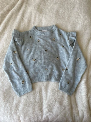 Ljusblå stickad tröja med volang - Supersöt ljusblå stickad tröja med små broderade blommor i vitt och gult. Tröjan har volangdetaljer på axlarna, rund halsringning och ribbade muddar vid ärmslut och nederkant. Perfekt för dig som gillar gulliga detaljer och en mjuk känsla.