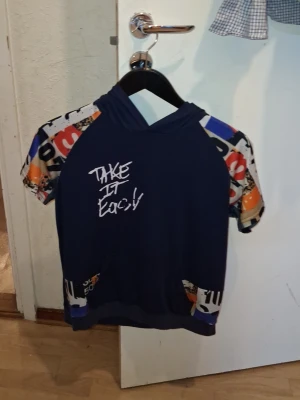 Blå hoodie med tryck och färgglada ärmar - Kortärmad hoodie i mörkblått med färgglada, grafiskt mönstrade ärmar och fickor. På bröstet står det 'Take It Easy' i vitt tryck. Huvan är enkel och materialet ser ut att vara mjuk bomullsblandning. Perfekt för en avslappnad streetwear-look.