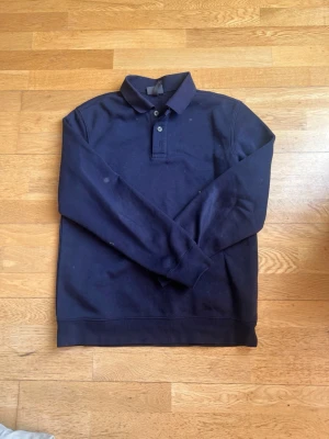 Mörkblå pikétröja med lång ärm - En stilren mörkblå pikétröja med lång ärm och klassisk krage. Tröjan har två knappar vid halsen och ribbade muddar vid ärmslut och nederkant. Perfekt för en avslappnad men ändå uppklädd look.