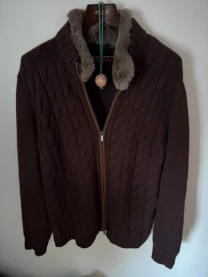 Cavour zip - Mysig brun kabelstickad kofta med dragkedja framtill och krage fodrad med mjuk päls. Långärmad modell med ribbade muddar och klassiskt mönster. Perfekt för kyliga dagar och ger en trendig look. Lite nopprig men de går lätt bort 
